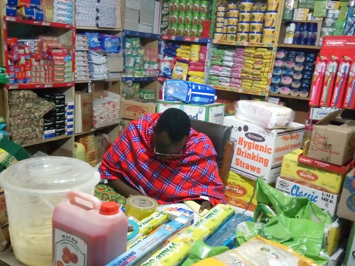 A shopkeeper in a red shuka sitting behind shelves of packaged goods